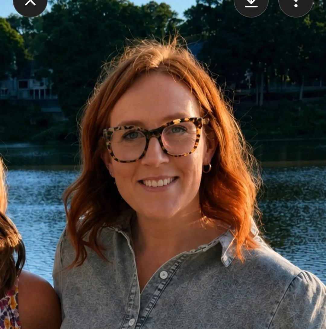 A woman with red hair, wearing leopard print glasses and a gray shirt, smiling outdoors near a body of water with trees in the background.
