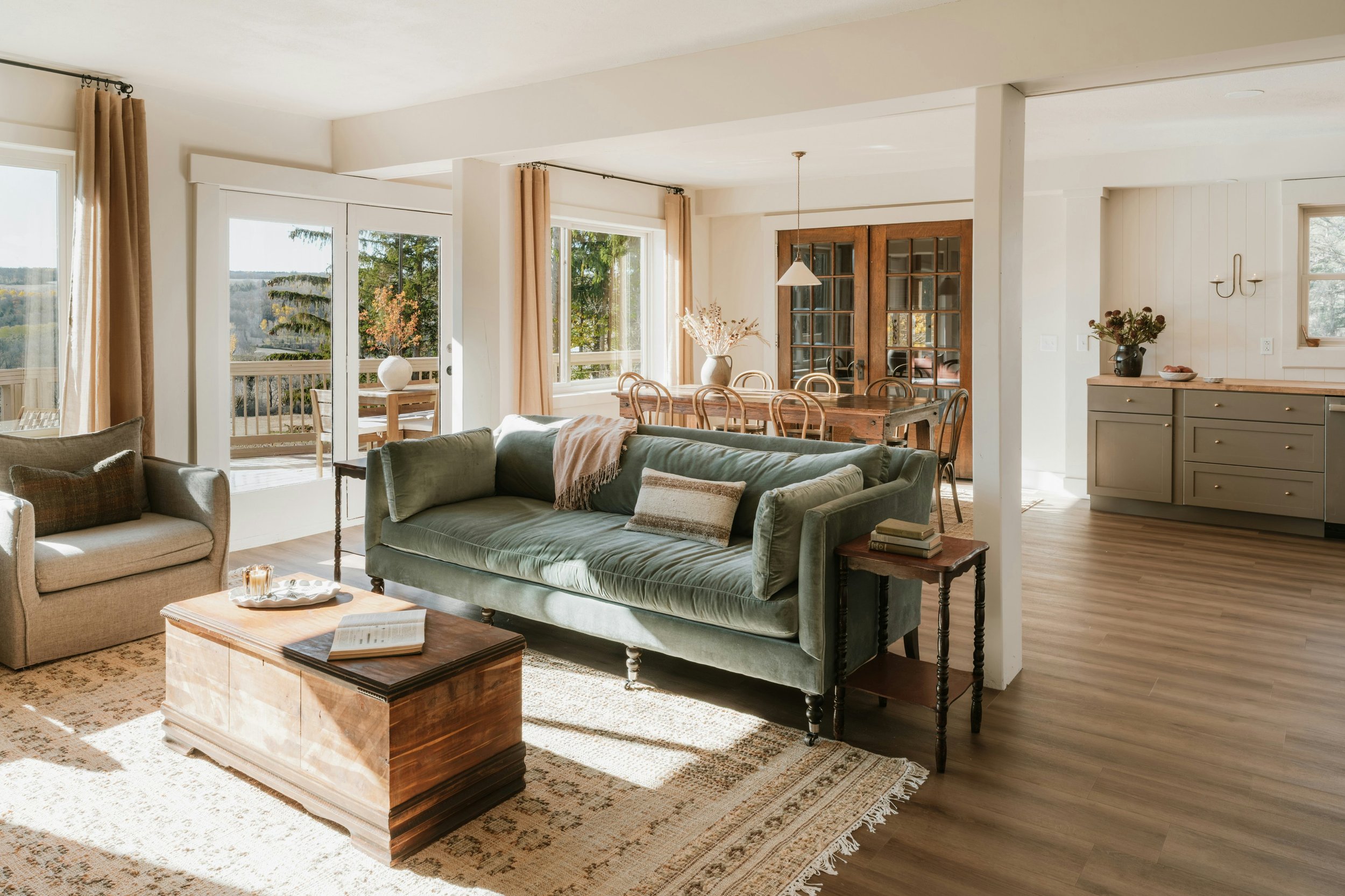 Bright living room with large window and door leading to a balcony, featuring a couch, armchair, wooden coffee table, and a view of trees outside.