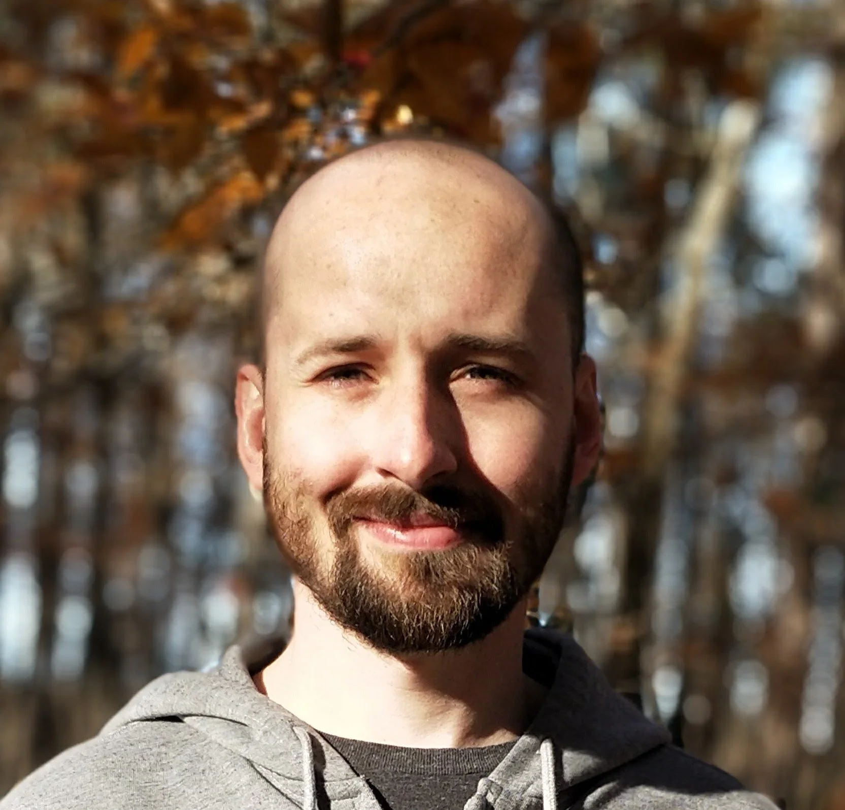 Close-up photo of a smiling man with a beard and mustache, bald head, wearing a gray hoodie, standing outdoors with a background of brown autumn leaves on trees.