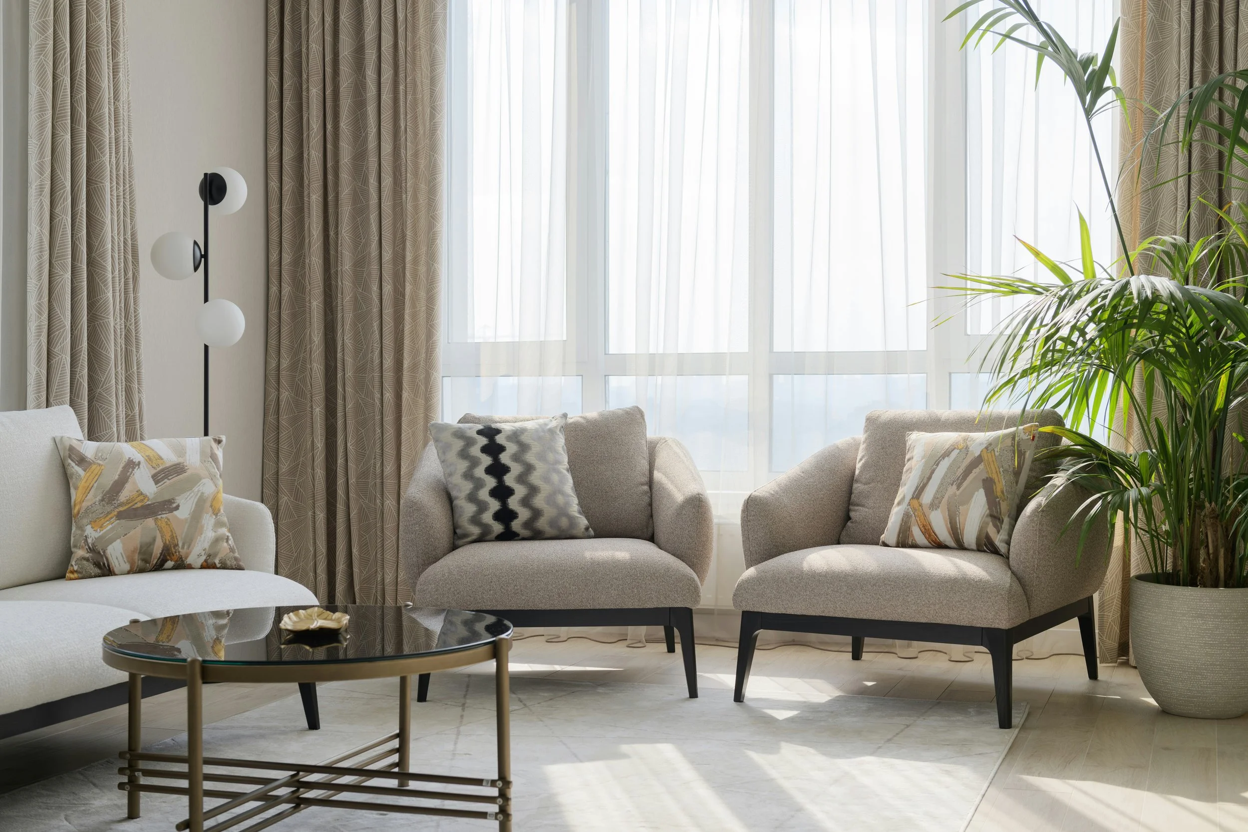 Living room with beige sofas, patterned pillows, large windows with sheer curtains, green potted plant, black coffee table, and a tall floor lamp with three globe-shaped bulbs.