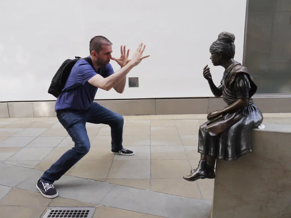 A man crouching and gesturing as if talking to a bronze sculpture of a seated woman.