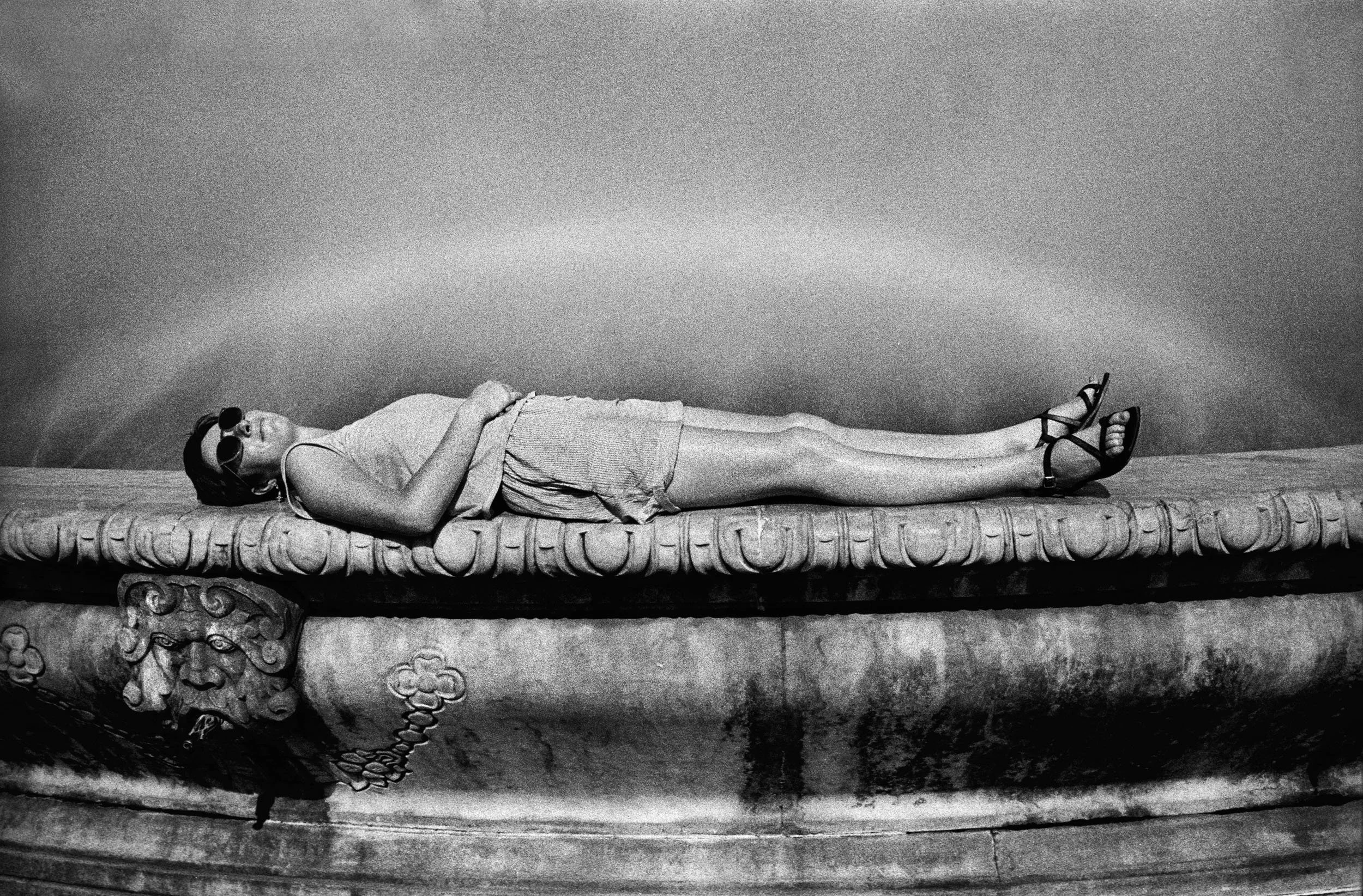 A woman lying on her back on an ornate ancient stone or marble platform, wearing sunglasses, a sleeveless top, a skirt, and high-heeled shoes, with her arms crossed over her stomach, against a plain background.