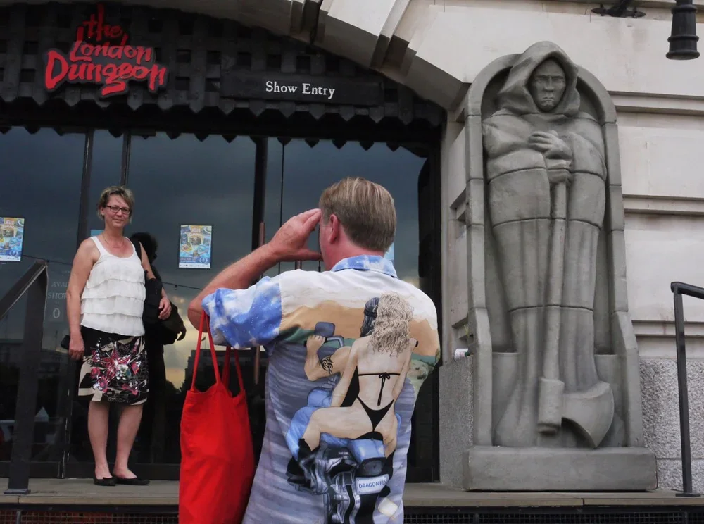 Man taking a photo of a woman in front of the entrance to The London Dungeon, which features a large stone sculpture of a stern-faced figure.