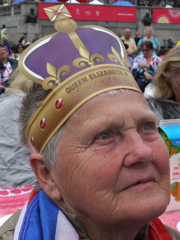 Elderly man wearing a purple and gold paper crown that says 'Queen Elizabeth II Since 1952' at an outdoor event, with people in the background.