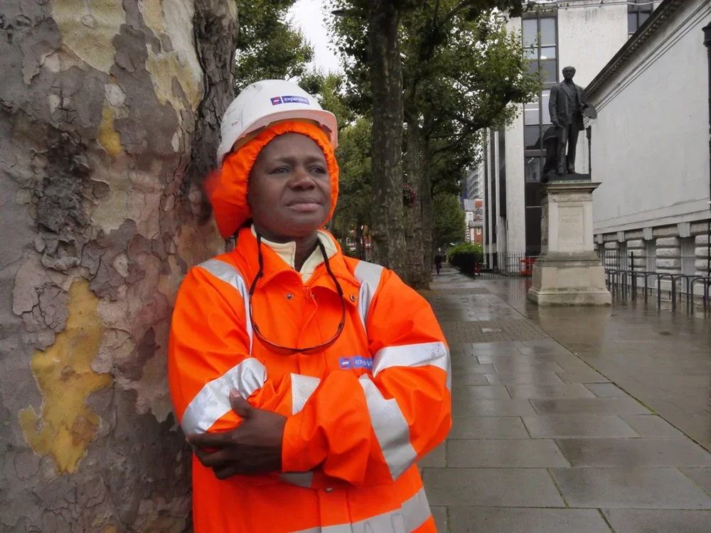 A woman in an orange safety jacket and white helmet stands on a city sidewalk, leaning against a large tree with a serious expression.