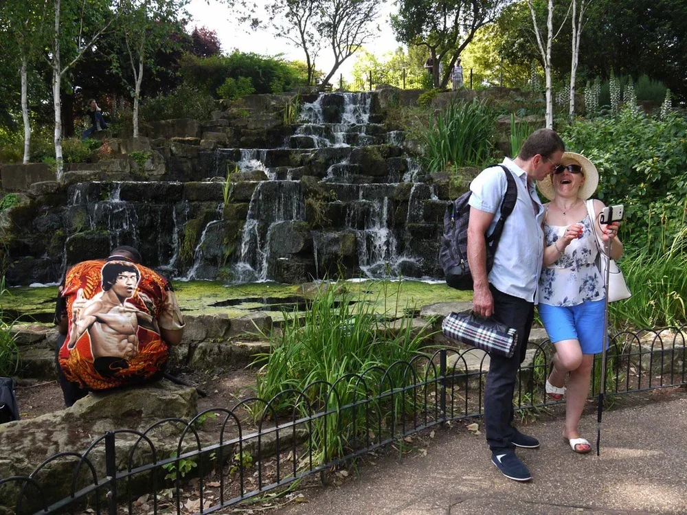 A waterfall in a park with three people; one person with a backpack and another holding a selfie stick, a shirtless man with an Asian actor's print on his shirt sitting nearby, and a woman wearing a large sun hat taking a selfie.
