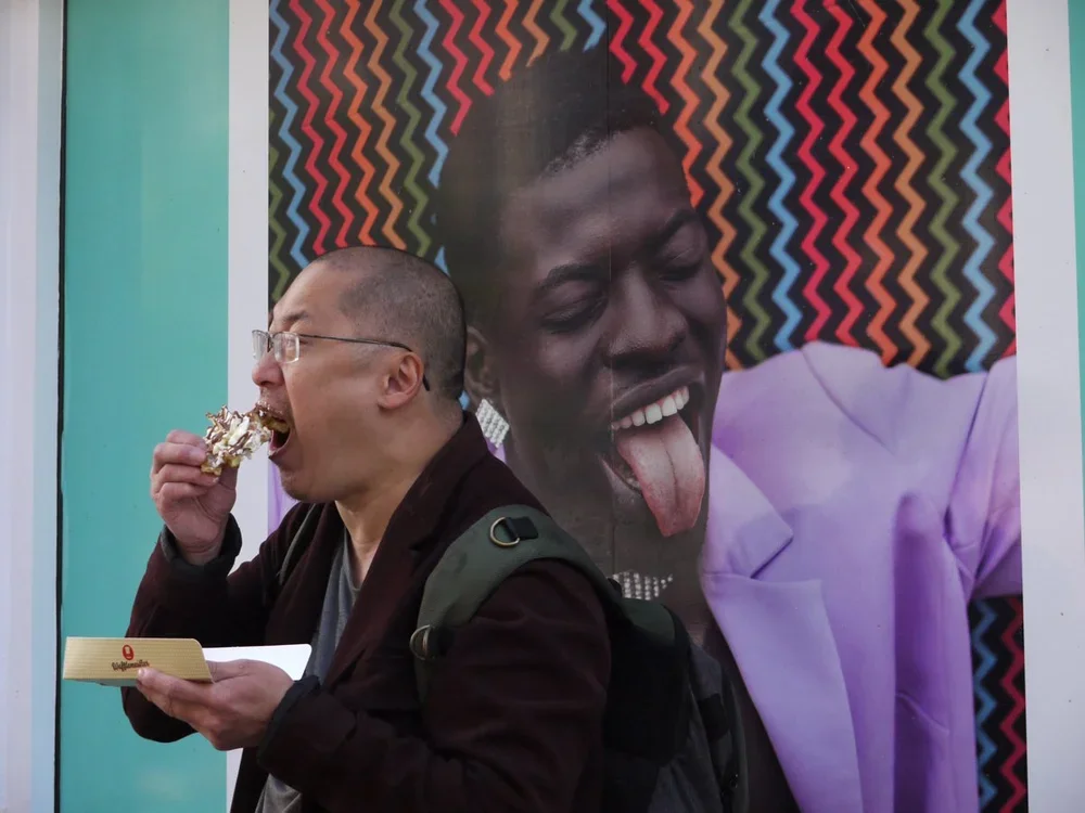 A person with glasses and a backpack eating ice cream stands in front of a large colorful poster of another person sticking out their tongue, who is dressed in a purple jacket and earrings. The poster features a multicolored zigzag background.