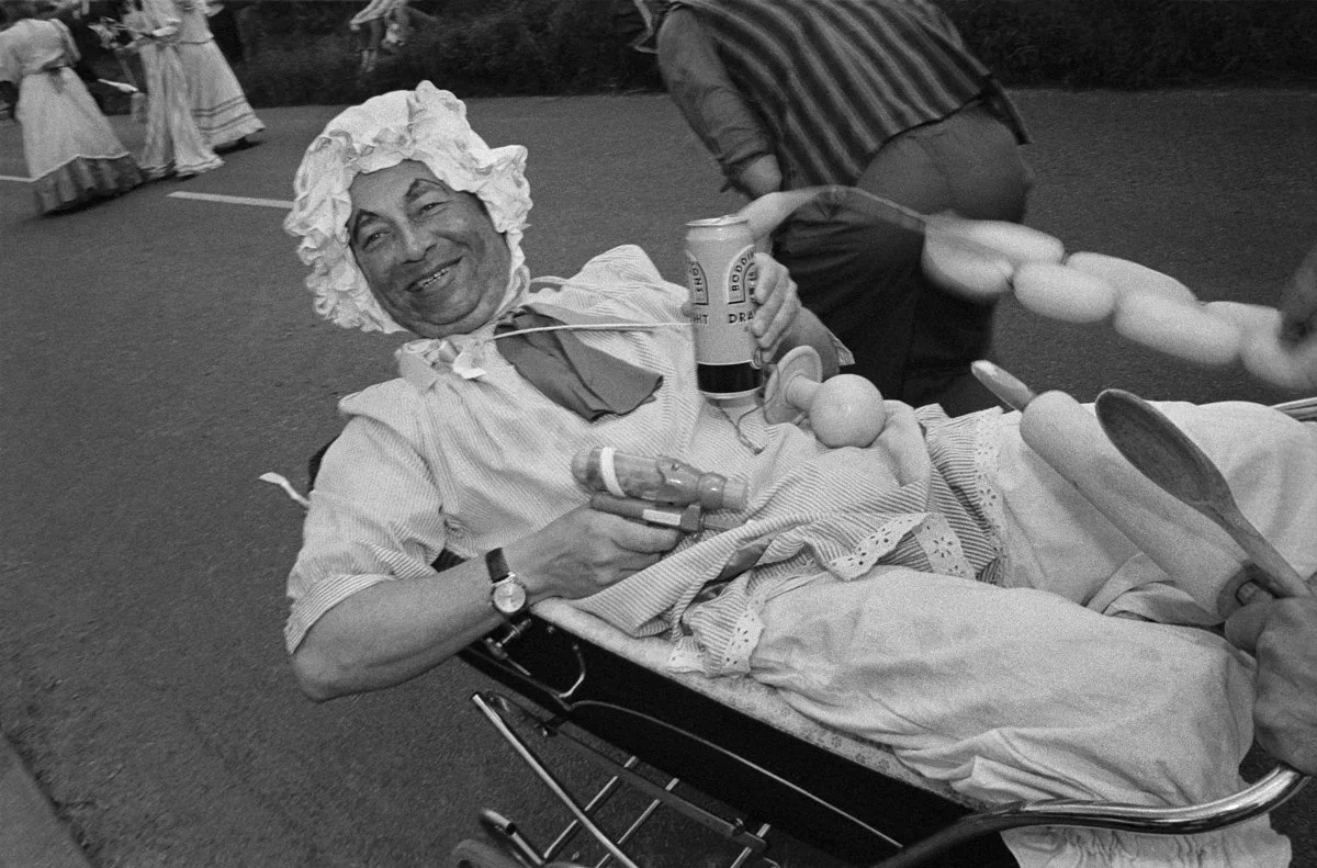 A person in vintage clown or entertainer costume lying on a stretcher, smiling and holding a can of Budweiser beer, at a festive outdoor event with people in period clothing in the background.