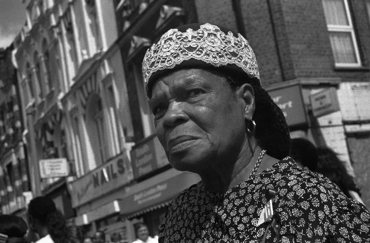 A black-and-white photo of an older woman wearing a lace headscarf and a patterned blouse, standing outdoors in an urban area with buildings and a crowd in the background.