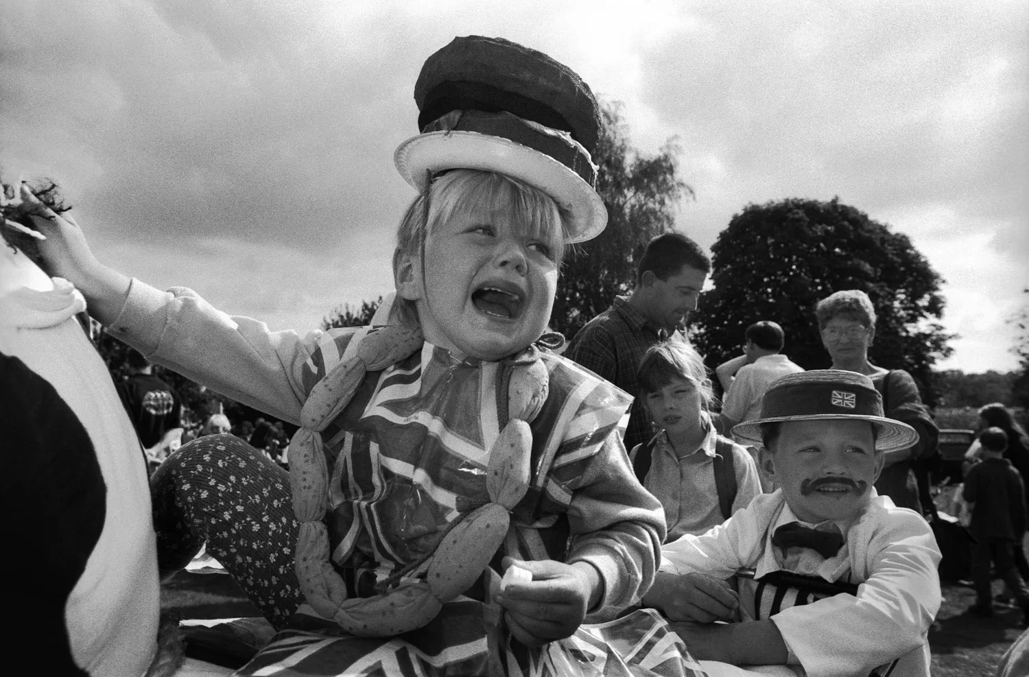 A black-and-white photo of children at a festival, dressed in clown and performer costumes, with one child in the center crying and wearing a hat with stacked cakes on it.