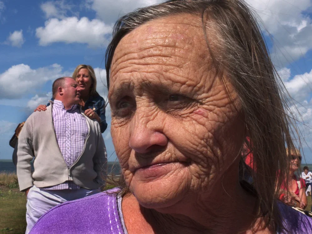 Close-up of an elderly woman with wrinkles, dark hair, and wearing a purple shirt, outdoors on a partly cloudy day.