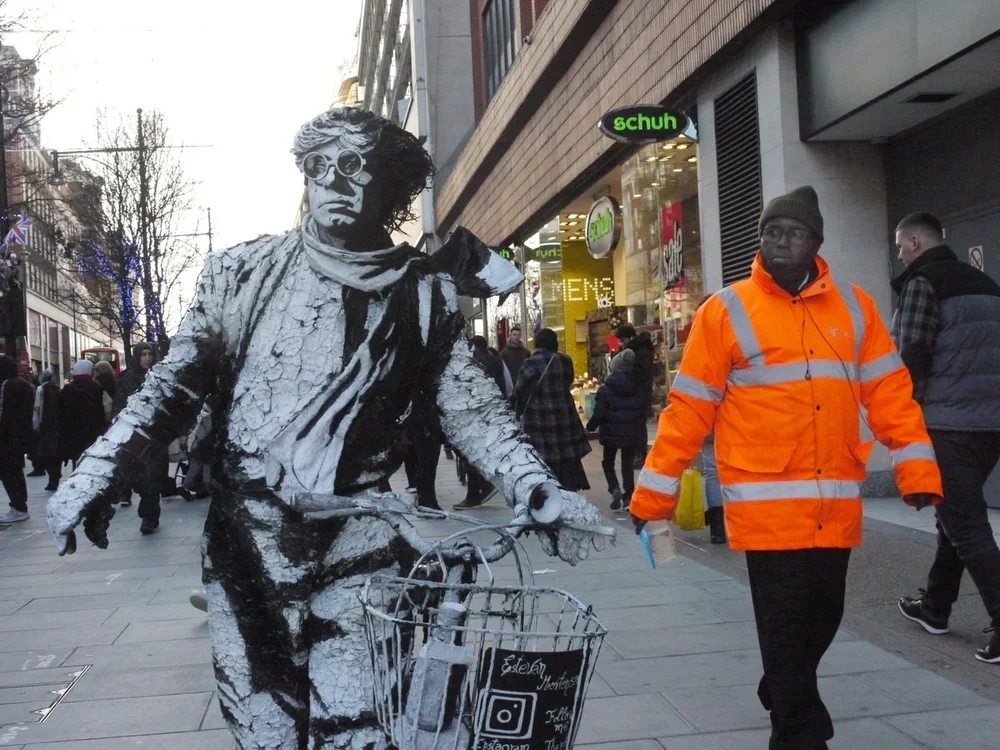A street performer dressed as a statue in monochrome costume, riding a bicycle with a basket, surrounded by pedestrians. The performer is painted in a black and white pattern, with sunglasses and a wig, in an urban shopping district.