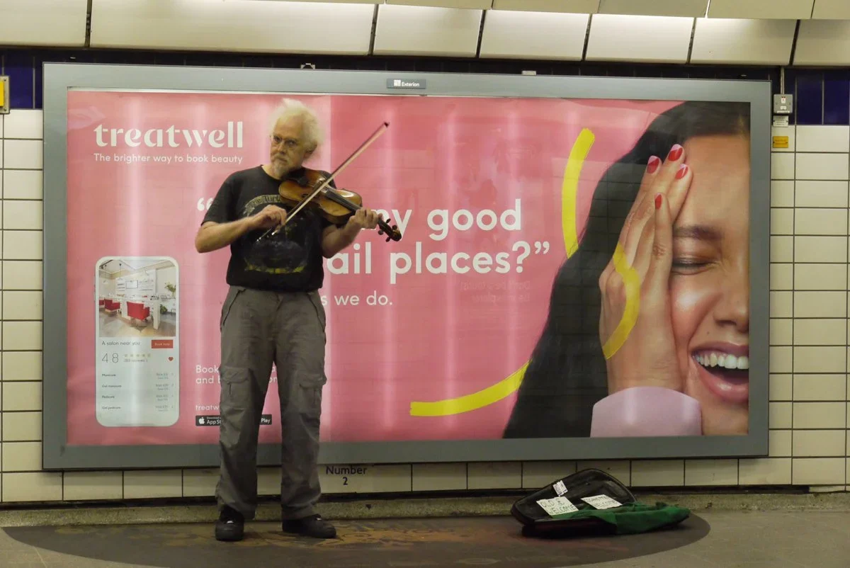 A man playing violin on a subway platform in front of a pink advertisement for Treatywell, a hotel booking app. The man has white hair, glasses, and is wearing a black t-shirt and gray pants. There is a violin case with a few items on the ground besi