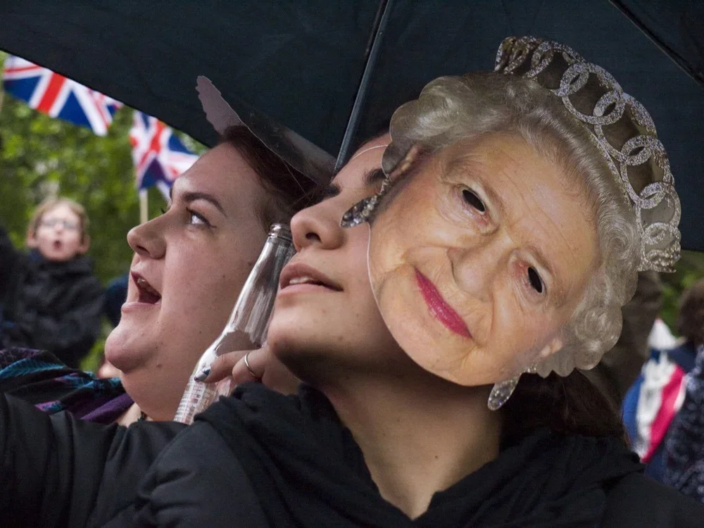 People at a public gathering under the rain, some holding umbrellas, one person wearing a mask of Queen Elizabeth II's face.