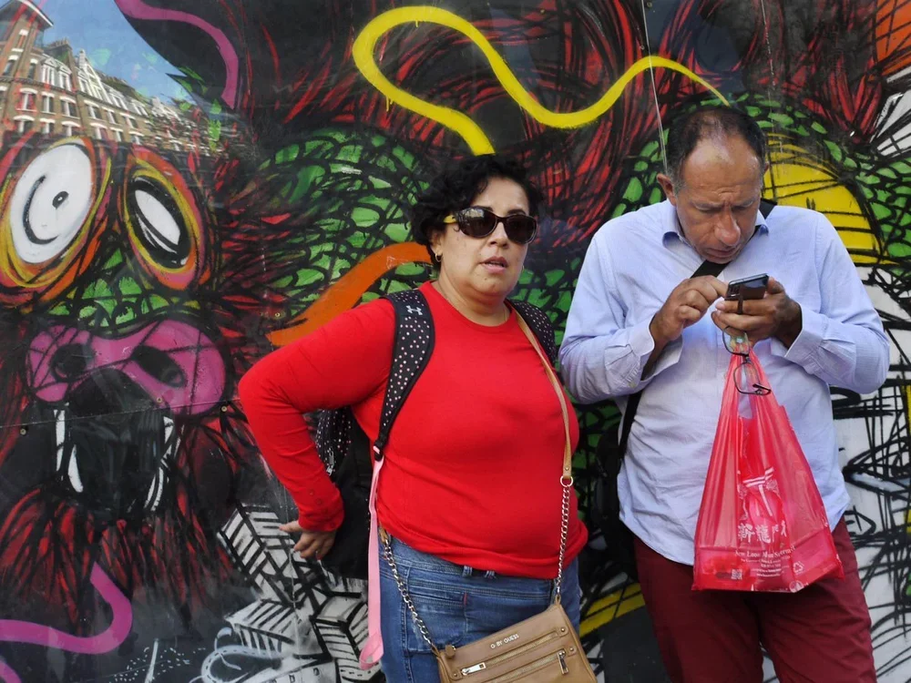 A woman in sunglasses and red shirt standing next to a man in a light blue shirt, looking at his phone, against a colorful graffiti wall with a dog face and abstract designs.