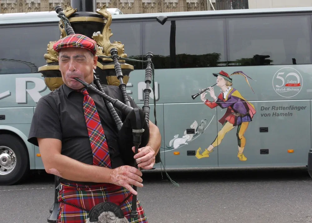 A man dressed in traditional Scottish attire playing bagpipes, standing in front of a bus with a cartoon image of a woman playing a flute and mice chasing her.