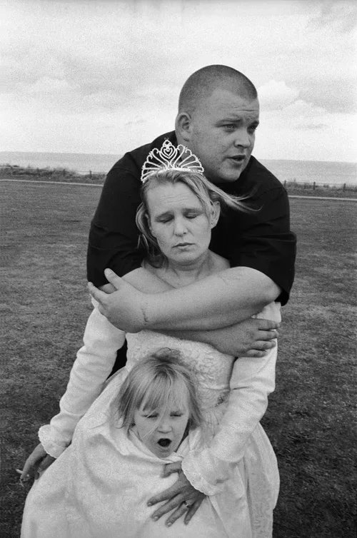 A young woman wearing a tiara is holding two children outdoors in an open field, with a big brother embracing her from behind. The young girl in front appears to be upset or crying, while the young woman and the boy behind her are looking distressed.