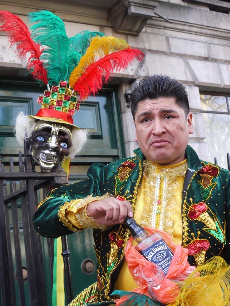Man wearing a colorful, sequined, green and yellow jacket with gold accents, holding a Jack Daniel's bottle, on a city street with a decorative mask and feathered headdress in the background.