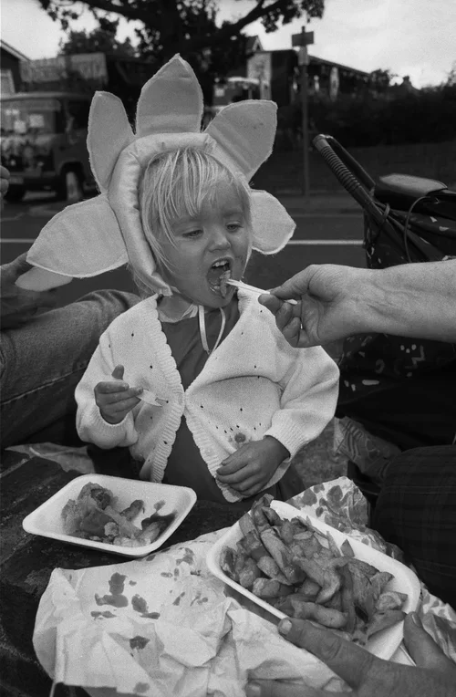 Child dressed as a flower receiving food from an adult, seated outdoors with containers of fries and sauce visible.