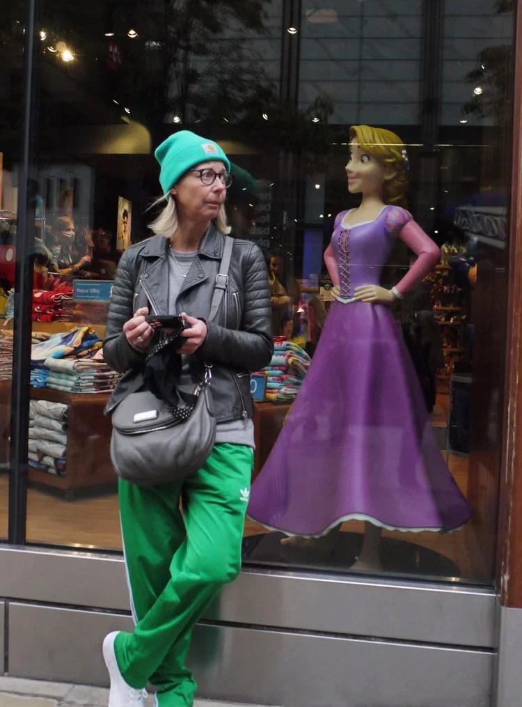 A woman standing outside a store window, looking at a cartoon princess figure in a purple dress displayed inside. The woman is wearing a teal beanie, glasses, a gray leather jacket, and green sweatpants. She is holding a phone in her hands and has a 