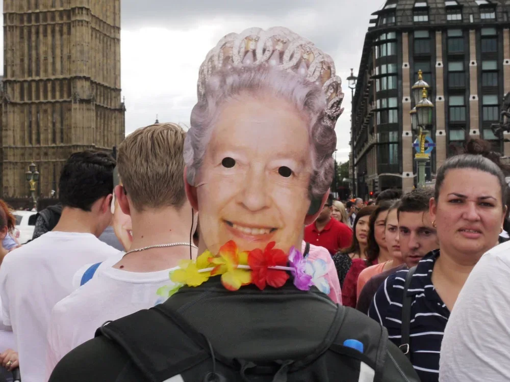 A person is wearing a mask with the face of Queen Elizabeth II, surrounded by other people in an outdoor urban setting.