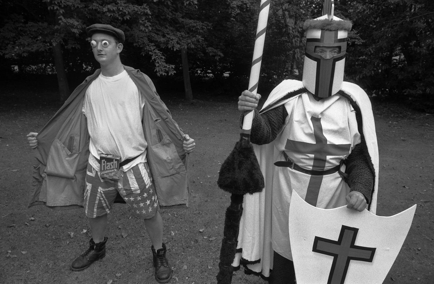 Two individuals in costume standing outdoors with trees in the background. The person on the left is wearing googly eye glasses, a cap, and casual clothing with a packet of Flash powder hanging from their belt. The person on the right is dressed as a