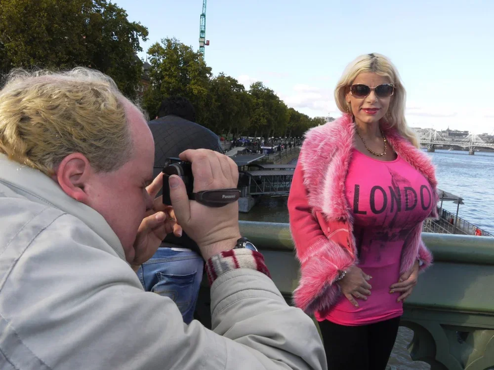 A woman in sunglasses, pink fur coat, and pink London shirt poses for a photograph on a bridge by a river, while a man with blonde hair takes her picture with a digital camera.