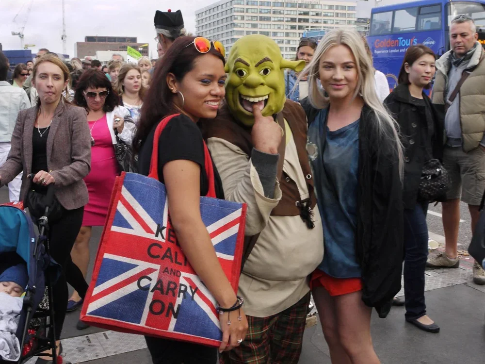 Three women, one dressed as Shrek, posing together at a crowded outdoor event with other people and buses in the background.
