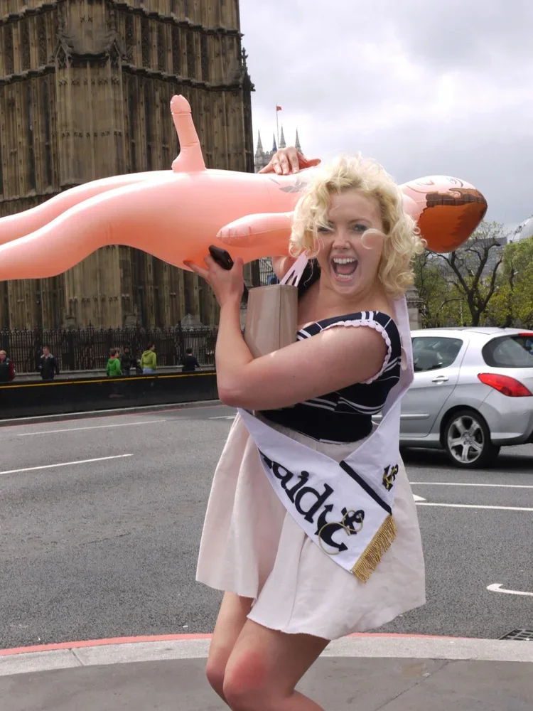 A woman with blonde curly hair smiling and laughing, holding a large pink inflatable mermaid, standing on a city street in front of a historic building with gothic architecture, cars parked nearby, and a cloudy sky.