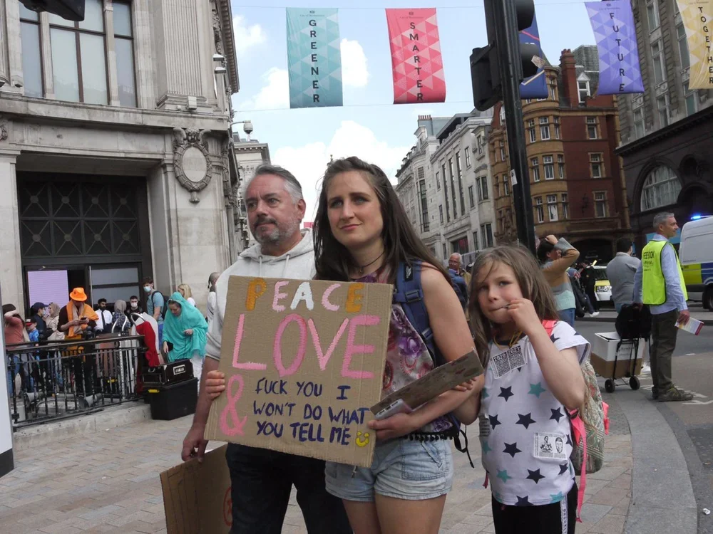 Young woman holding a sign that says "Peace, Love, &" with additional handwritten messages, standing on a city street with a man and a young girl, surrounded by people at a public event.