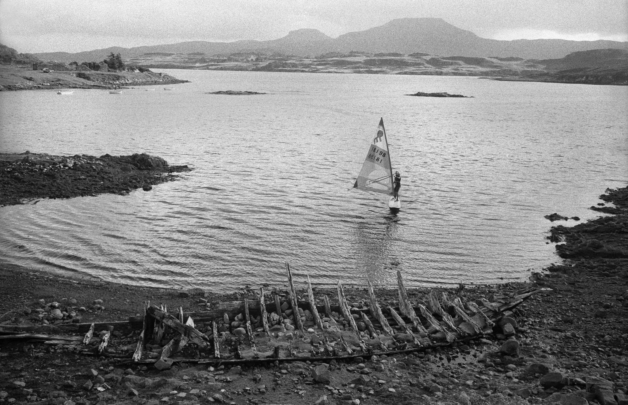 A black-and-white photo of a coastal scene with a person windsurfing on the water, rocky shoreline, and distant hills in the background.
