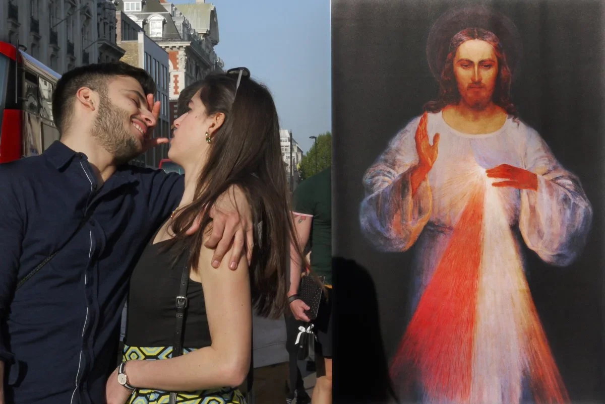 A young couple with light skin standing close, smiling, and touching foreheads outdoors in a city street with historic buildings. To the right, a religious painting of Jesus Christ with a halo, raising his right hand in blessing and pointing to his h