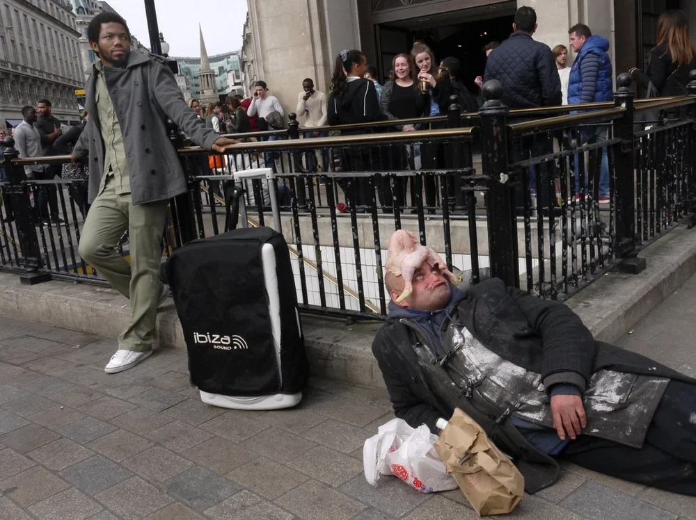 A street performer dressed as a horror movie character lying on the sidewalk with makeup and prosthetics, while a man stands nearby with a large portable speaker and a suitcase, with a crowd of people gathered outside in the background.