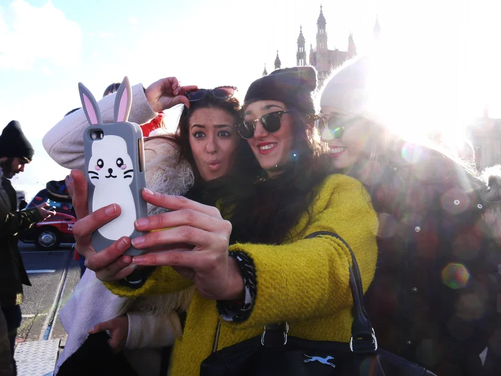Three women taking a selfie outdoors with a historic building in the background during a sunny day.
