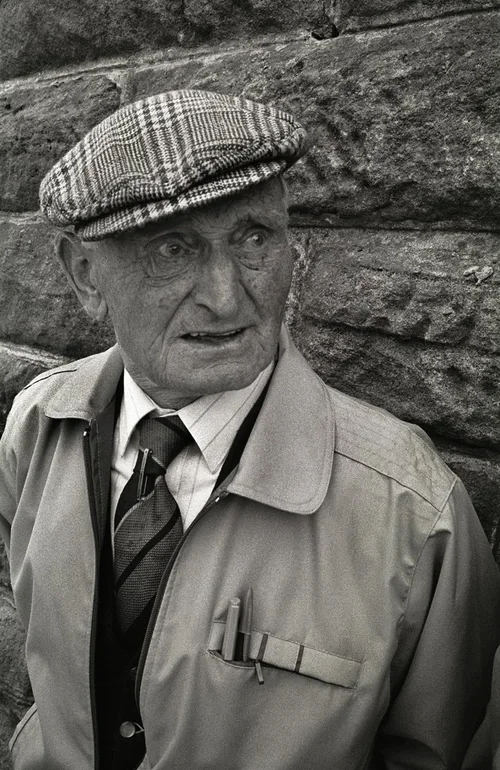 An elderly man wearing a plaid flat cap, a light-colored jacket, and a striped tie, standing against a stone wall, looking to the side.