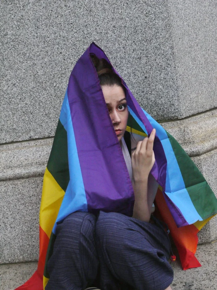 Person sitting against a stone wall, partially wrapped in a rainbow pride flag, looking at the camera with a serious expression.