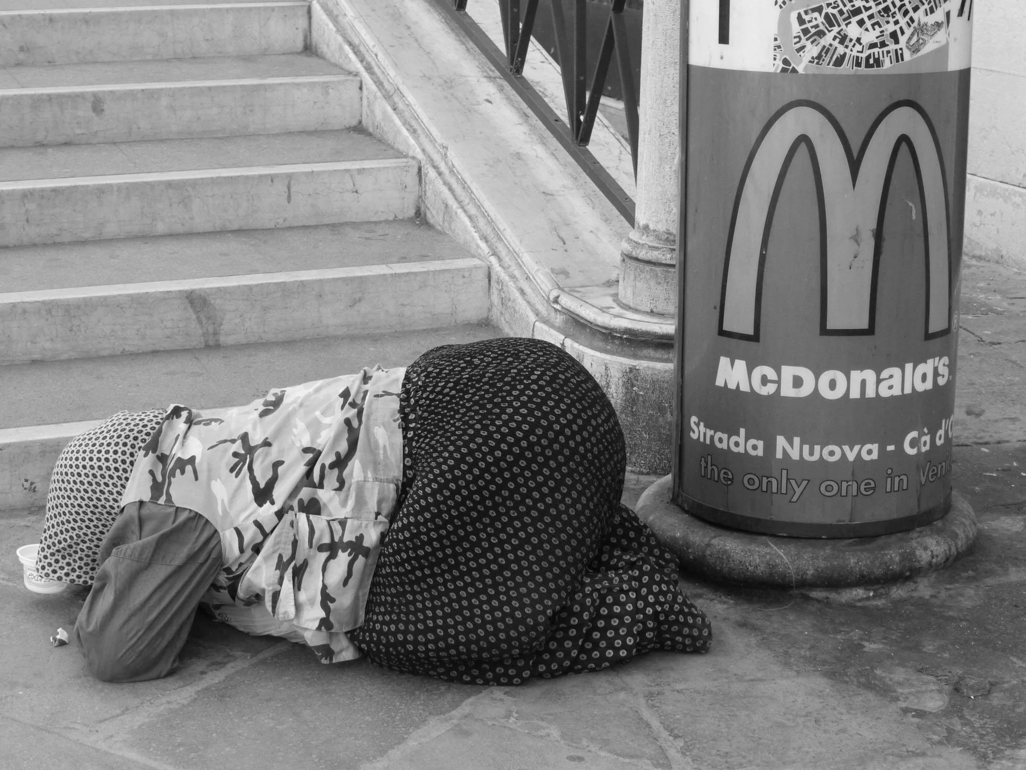 A person with curly hair lying on the ground next to a McDonald's sign on a city sidewalk, near a staircase and a metal railing.