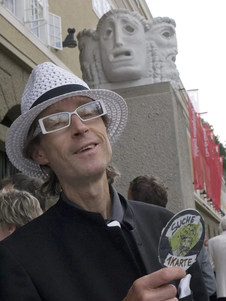 A man wearing a white sun hat, white sunglasses, and a black jacket is smiling and holding a small sign with a face and the text 'SUCHE AKARTE'. He is standing outdoors in front of a building with a large stone sculpture of three faces on top.