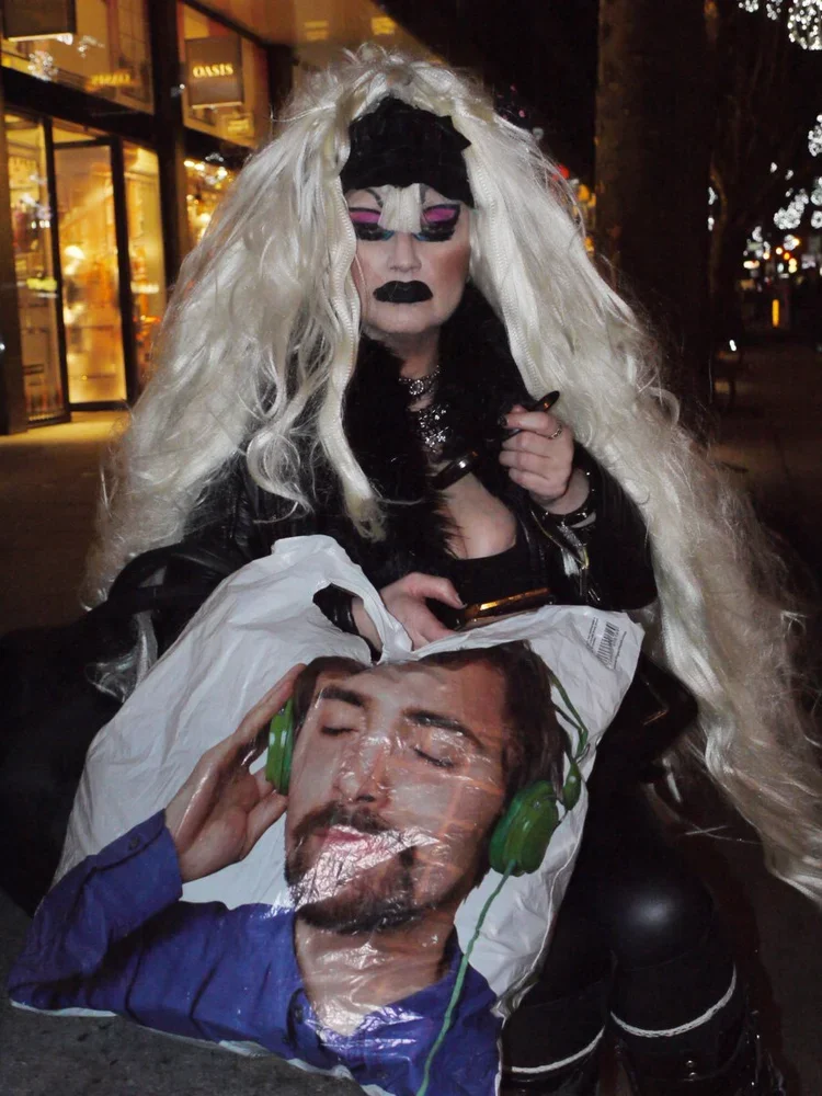 Person with long, curly platinum blonde hair, dramatic makeup with pink eyeshadow and black lipstick, wearing a black jacket, holding a shopping bag with a printed face of a man wearing green headphones.