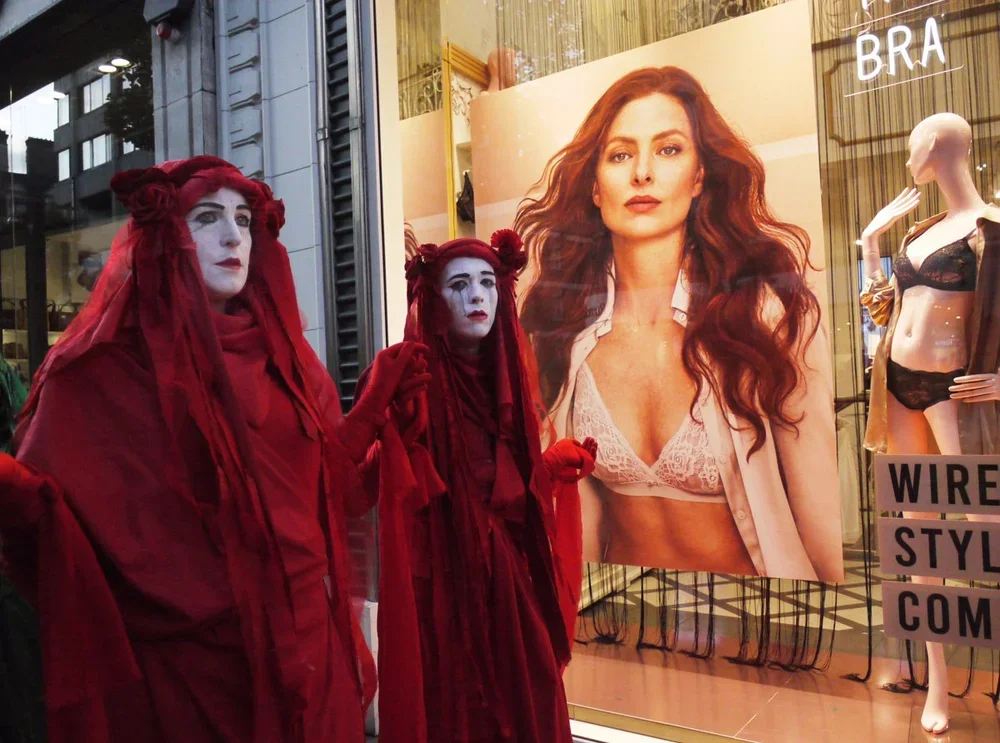 Two mannequins dressed as The Little Mermaid in red costumes with white face paint in a store window display. Behind them, there is a large poster of a woman with long, wavy red hair wearing a white lace bra and an open shirt. To the right, a mannequ