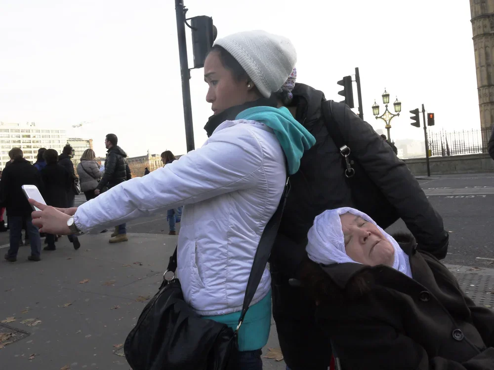 A woman in a white jacket and gray beanie is taking a selfie while pushing a wheelchair with an elderly woman in a white headscarf on a city street, with other pedestrians and historic architecture in the background.