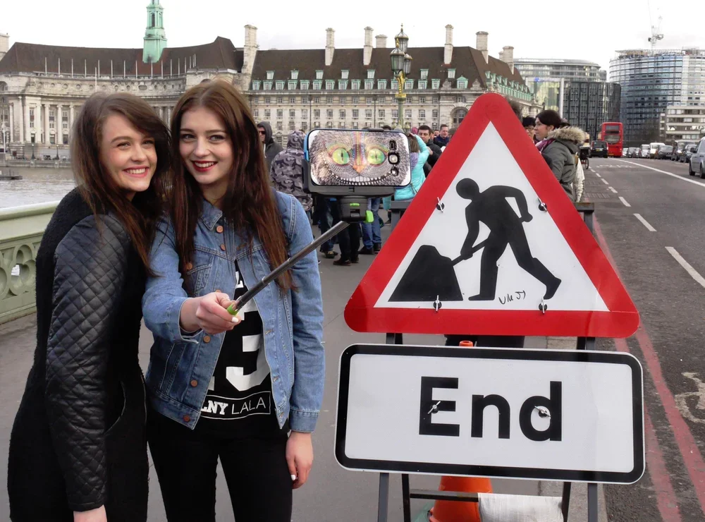 Two smiling women taking a selfie with a selfie stick on a city bridge, with a street sign reading 'End' and a construction sign behind them.