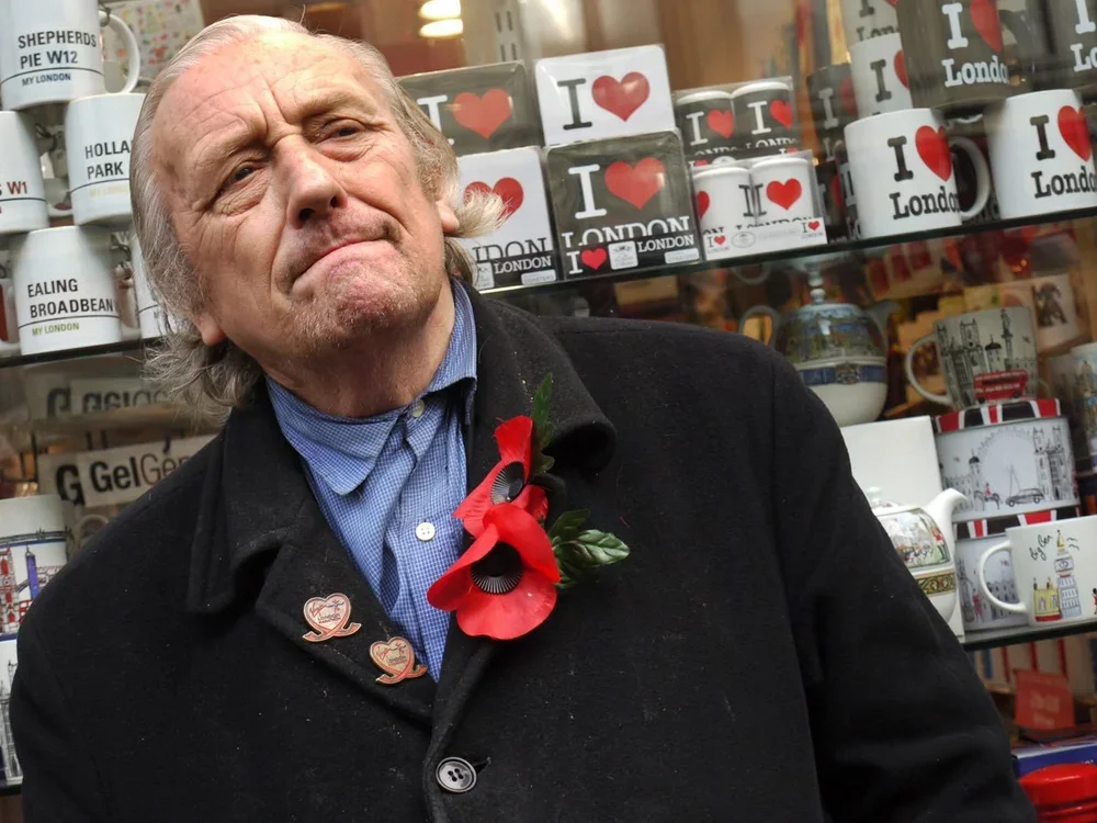 An elderly man with gray hair, wearing a black coat, blue shirt, and a red poppy pin, stands outside a shop window filled with mugs that have 