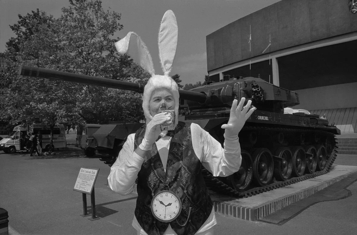 A person in a bunny costume with ears, holding a drink in one hand and gesturing with the other, standing in front of a tank on display.