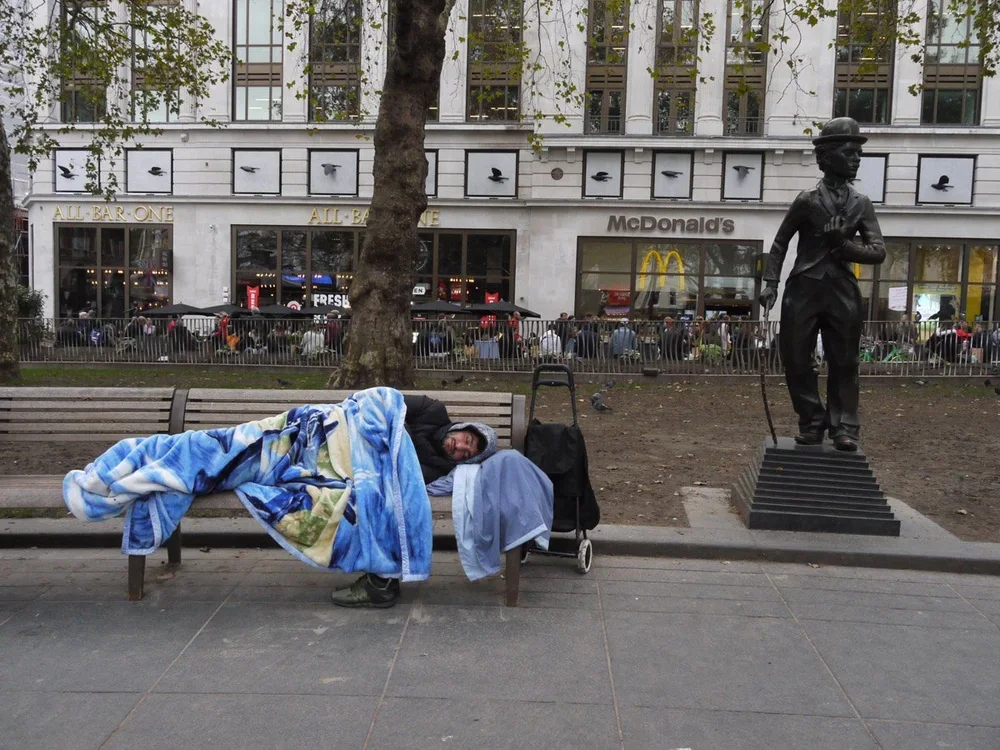 A man sleeping on a park bench with a blanket, next to a stroller, in front of a McDonald's restaurant and a statue of a soldier in an urban setting.