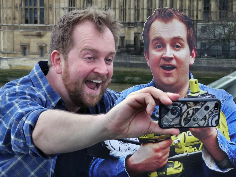 Two men are taking a selfie together outdoors, with a historic building in the background. One man has a beard and is wearing a blue plaid shirt, and the other man has red hair, wearing a blue shirt. The person with the beard is holding the phone cam
