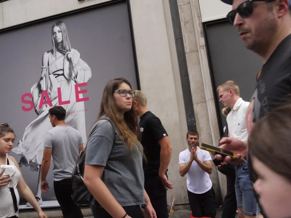 A group of people standing and walking on a city street sidewalk in front of a store with a sale advertisement poster featuring a model in a dress.