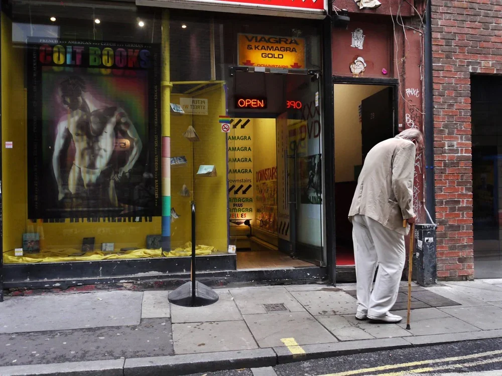 An elderly man with a cane standing on a city sidewalk outside a store with open door. The store has psychedelic artwork of a muscular man in a diabetic coma on the window, and signs for Viagra and Kamagra inside. There is a neon sign that reads 'OPE