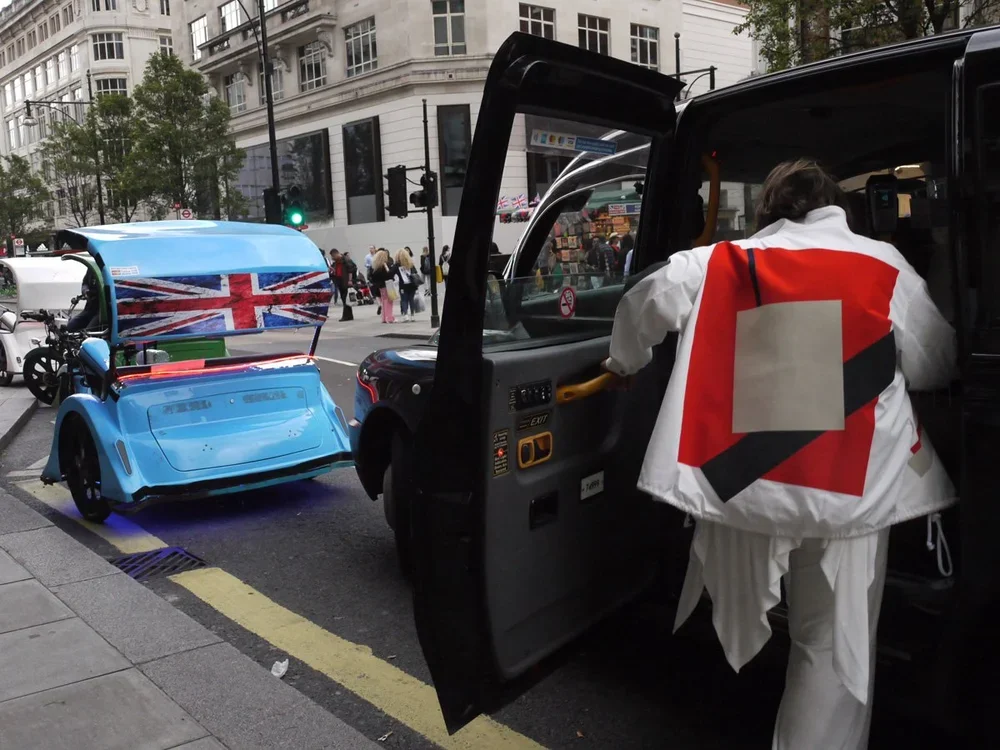 A person wearing a jacket with a red, white, and black design is stepping into a black Uber vehicle on a city street. A small, blue electric vehicle with a Union Jack design on its back is parked nearby.