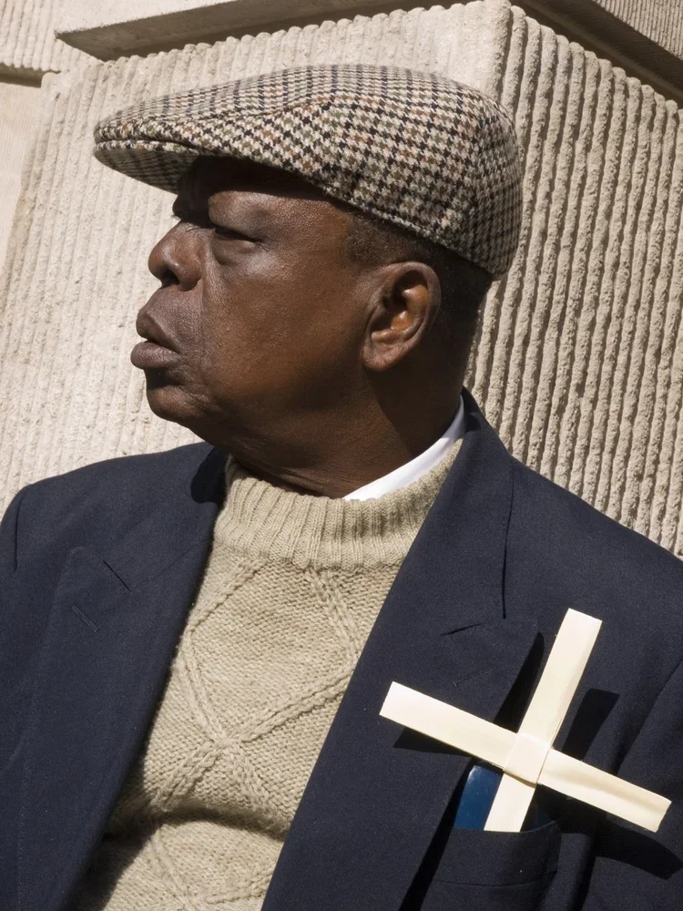 A man wearing a checkered flat cap, a beige knitted sweater, a dark blazer with a white shirt underneath, and holding a white cross, standing against a textured beige wall.